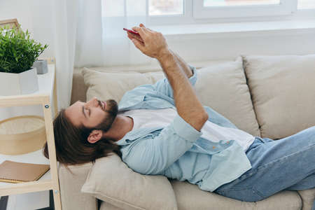 A man with a beard lies on the couch during the day at home and looks at his phone relaxing on his day off, a man gambling on the stock market online on his phoneの写真素材
