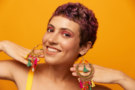 Portrait of a young woman with a short haircut and colored hair smiling and showing her tongue at the camera on an orange background with earrings accessories in the studioの写真素材