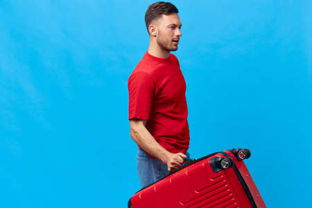 Cheerful confident tanned handsome man in red t-shirt going with suitcase ready for vacation trip posing isolated on blue studio background. Copy space Banner Mockup. Trip journeys conceptの写真素材