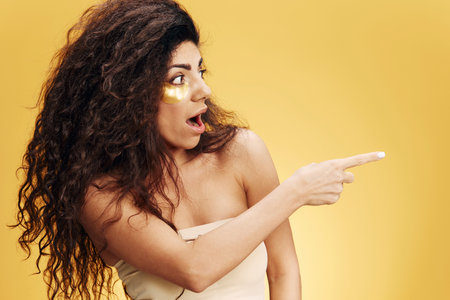 Shocked curly pretty Latin female with gold hydrogel patches under eye point finger look aside posing isolated over yellow background. Cosmetic product ad Natural beauty concept Studio profileの写真素材