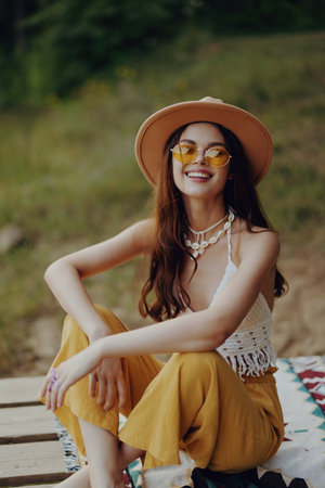 A beautiful young woman in a hippie look with yellow pants, knit top, hat, and yellow glasses sits on the ground on a plaid in the park and smiles happily looking at the cameraの写真素材