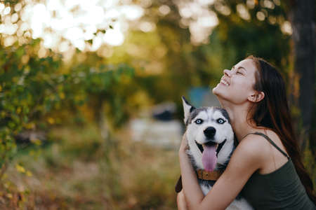 A woman with a husky breed dog smiles and affectionately strokes her beloved dog while walking in nature in the park in autumn against the backdrop of sunsetの写真素材