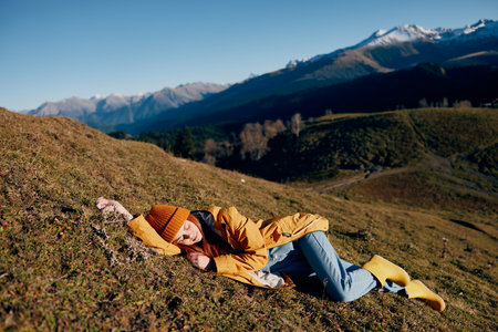 Woman lying on a hill on the grass rest smile with teeth looking at the mountains in the snow in winter in a yellow raincoat and jeans happy sunset trip on a hike, freedom lifestyleの写真素材