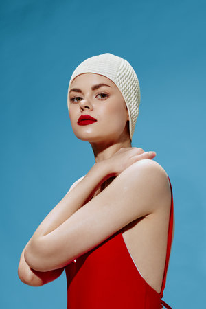 An elegant spectacular swimmer in a red swimsuit swimming cap touches her neck posing on a blue backgroundの写真素材