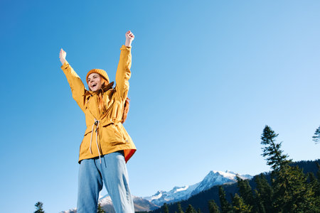 Woman with red hair hiker in yellow raincoat and cap standing on the mountain hands up happiness with a view of snowy mountains travel in winter and hiking in the mountains in the sunset freedomの写真素材