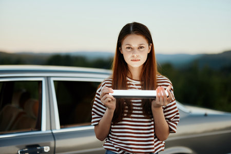 A woman holds a flashlight standing by her car on a nature trip to a campground and smiles. Travel...