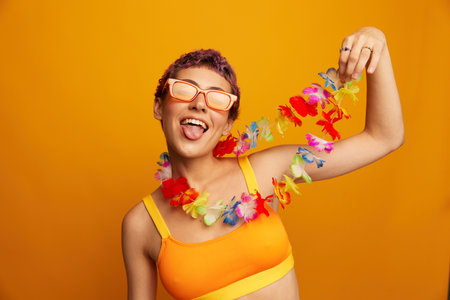 Portrait of a woman with a floral Hawaiian wreath around her neck is having fun dancing and smiling in bright clothes on an orange background wearing sunglasses, lifestyle party Hawaiian styleの写真素材