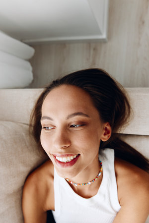 A beautiful young woman lies at home on the couch close-up on a wide-angle lens in a relaxed pose in a white tank top and blue jeans. Lifestyle in comfortの写真素材