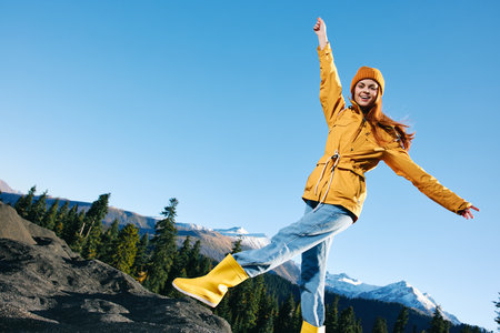 Woman with red hair hiker jumping on mountain hands up happiness overlooking snowy mountains in yellow raincoat and cap travel autumn and hiking in the mountains in the sunset freedomの写真素材