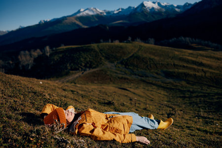 Woman beautifully lying on a hill recreation smile with teeth in the mountains in the autumn in a yellow raincoat and jeans happy sunset trip to hike the mountains in the snow, freedom lifestyleの写真素材