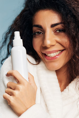 Cheerful curly Latin lady in bathrobe with toner spray in hand pose isolated on pastel blue background, feeling excited of effective skincare routine. Skincare concept Natural Beauty Cosmetic productの写真素材