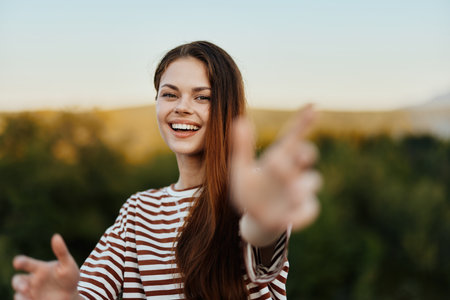 Woman smiling while looking at the camera and pulling her hands to the camera close-up in nature with a view of the mountains. Happy travel lifestyle follow meの写真素材