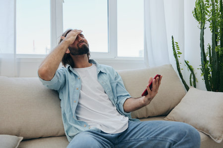 A young man looks angrily and sadly at the phone screen at home sitting on the couch. Bad news and...