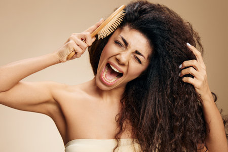 DAMAGED TANGLED HAIR CONCEPT. Screaming crying unhappy tanned awesome curly Latin lady with hairbrush pulling hair posing isolated on pastel beige background, closing eyes. Copy space, good offerの写真素材