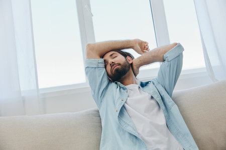 A man sits on the couch yawning and stretching after a nap, lack of sleep and fatigue from work and improper daily routine.の写真素材