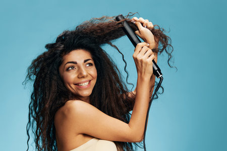 Cool offer. Beautiful Latin curly woman using hair straightener, looking aside, posing isolated on blue wall background. Hair routine concept, haircare, dry damaged hair ironing, hairdressing. Bannerの写真素材
