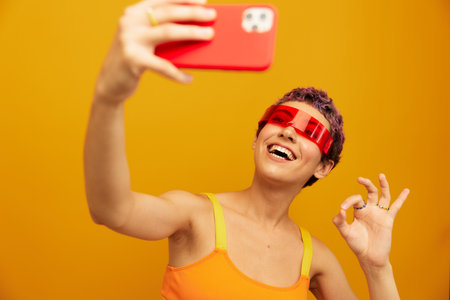 Woman smiles and shows her tongue in unusual glasses millennial takes selfies on her phone in sportswear against an orange studio backdrop, free spaceの写真素材