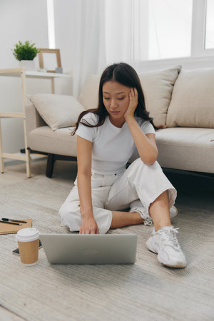 Young Asian female student looking at laptop screen sitting on floor at home with notebooks to write, lifestyle work freelanceの写真素材
