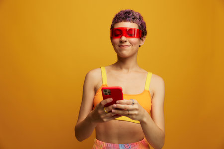 Woman blogger smiling in unusual red millennial glasses holding her phone and looking at the screen against an orange background in a stylish yellow topの写真素材