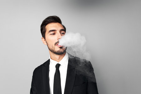 Man businessman in suit smokes e-cigarette and blows steam from his mouth, smoking and health stress in gray background close-up of face at wide camera angleの写真素材