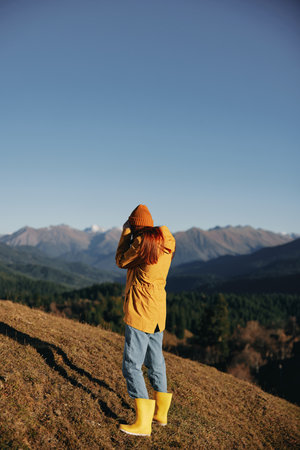 Woman walking full-length on a hill in the autumn and looking at the mountains in a yellow raincoat and jeans happy trip in the sun sunset hiking, freedom lifestyleの写真素材