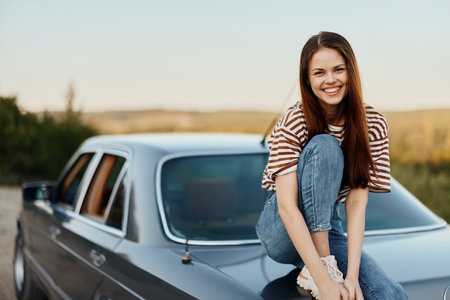 A young woman sits on the trunk of a car, laughs and rests after a difficult road and admires nature with a beautiful view. Stopping is also part of the journeyの写真素材