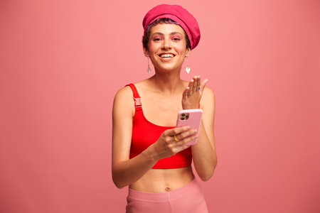 Young woman blogger with colored pink hair and short haircut flipping through the phone screen and typing a message with a smile and surprise in stylish clothes on a pink backgroundの写真素材