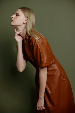 Fashion profile studio portrait of blonde stylish young woman in trendy leather trench coat posing isolated on olive green background. Resale offer for marketplace. Huge sale conceptの写真素材