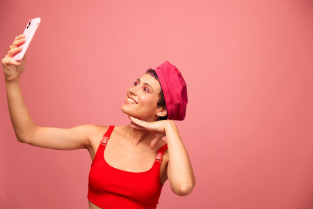 A young woman blogger with colored pink hair and a short haircut takes a picture of herself on the phone and broadcasts a smile in stylish clothes and a hat on a pink background monochrome styleの写真素材