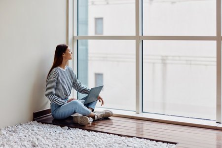 A woman freelancer sits with her laptop full-length at a large window, looking out at tall buildings...