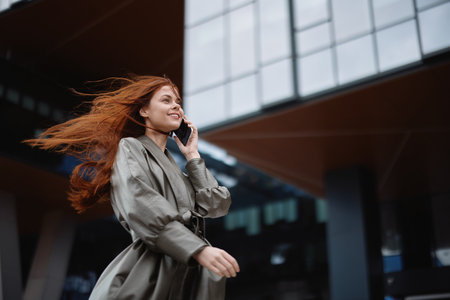 Woman walking around town and talking on the phone in trendy clothes against a backdrop of tall city buildings with an out-of-focus smile in windy weatherの写真素材