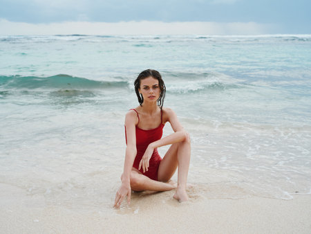 Woman with wet hair after swimming in the ocean in a red swimsuit sits on the beach in Bali and looks into the camera, a trip to Indonesiaの写真素材