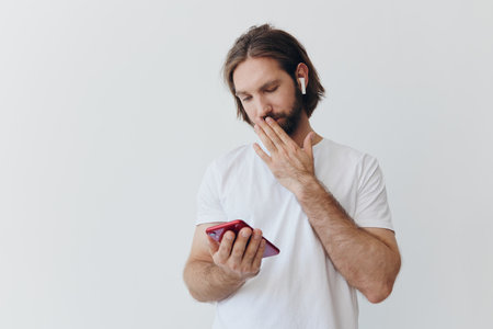 A man is holding a phone with wireless headphones in his ears and reading a social media message online freelancer job correspondence on a white backgroundの写真素材