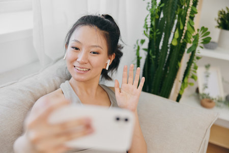Athletic Asian woman in yoga costume blogger filming herself on phone camera sitting on couch and smiling with headphones waving, freelance work from home, recording sports workout lifestyleの写真素材