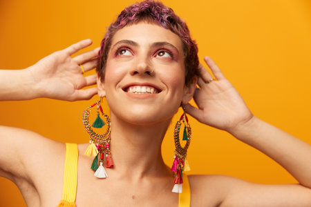Portrait of a young woman with a short haircut and colored hair smiling and showing her tongue at the camera on an orange background with earrings accessories in the studioの写真素材