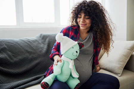 A pregnant woman is sitting on the couch with a childrens toy rabbit in her hands talking to the child for Easter. Lifestyle of a pregnant woman, preparation for childbirth, last month of pregnancyの写真素材