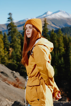 Woman in yellow raincoat with red hair on a hike stands in front of mountains in yellow cap autumn sunset lightの写真素材