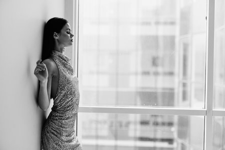 Black and white photo of a woman in a stylish sequined party dress posing near a window bwの写真素材