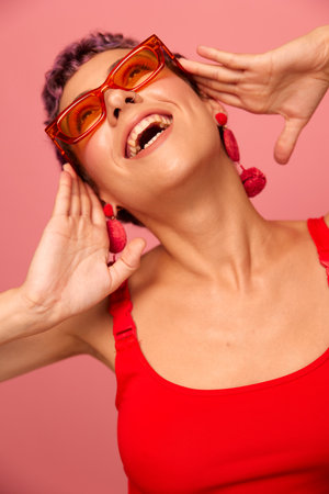 Young athletic woman with a short haircut and purple hair in a red top and pink yoga leggings in sunglasses with an athletic figure smiles and dances on a pink backgroundの写真素材