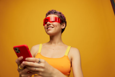 Woman blogger smiling in unusual red millennial glasses holding her phone and looking at the screen against an orange background in a stylish yellow topの写真素材