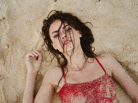 Woman with wet hair lies in the sand looking at the camera on the ocean in a swimsuit, dramatic portrait of a girl in the sand with a tanned bodyの写真素材