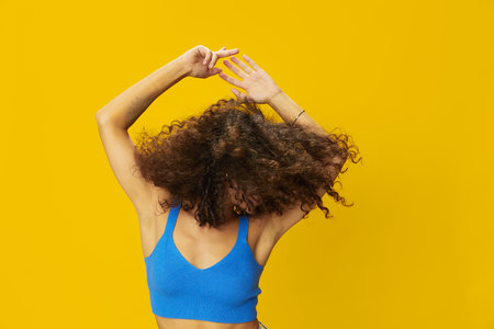 Woman with curly afro hair in a blue T-shirt on a yellow background dancing flying hair with sunglasses yellow, hand signs, look into the camera, smile with teeth and happiness, copy spaceの写真素材