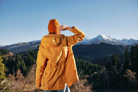 Autumn woman standing with her back up with her hands in the air hiking in the mountains and happiness in a yellow cape full-length standing in front of the trees and mountains in the sunset, freedomの写真素材
