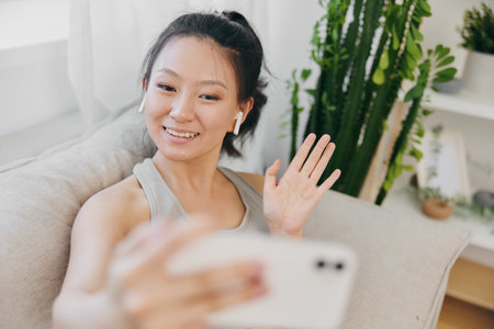 Athletic Asian woman in yoga costume blogger filming herself on phone camera sitting on couch and smiling with headphones waving, freelance work from home, recording sports workout lifestyleの写真素材