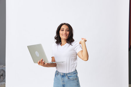 A woman freelancer with a laptop in her hands on a white background in a white t-shirt raised her hand up, emotion of emotion and smile, copy spaceの写真素材