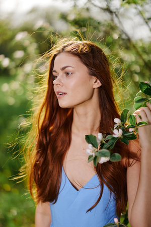 Beautiful woman smile with teeth with long red hair near a tree in the summer sun in nature in the park smiling without allergies in a blue dress, the concept of health and beautyの写真素材