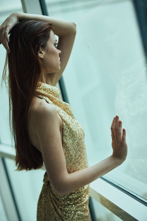 Beautiful fashion woman in stylish expensive gold sequined dress poses looking out the window at the city in a hotel, Christmas partyの写真素材