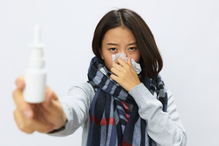 Asian woman flu cold with stuffy nose holds allergy nasal spray with sore throat on white backgroundの写真素材