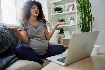 Pregnant woman smile sits at home on the couch with a laptop and talks with a doctor on video chat, works online. Lifestyle of a pregnant woman, preparation for childbirth, last month of pregnancyの写真素材