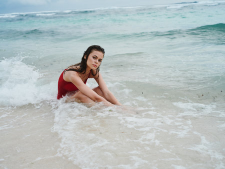 A woman in a red swimsuit sits on the sand by the ocean in the waves on the beach of Bali Island and smilesの写真素材
